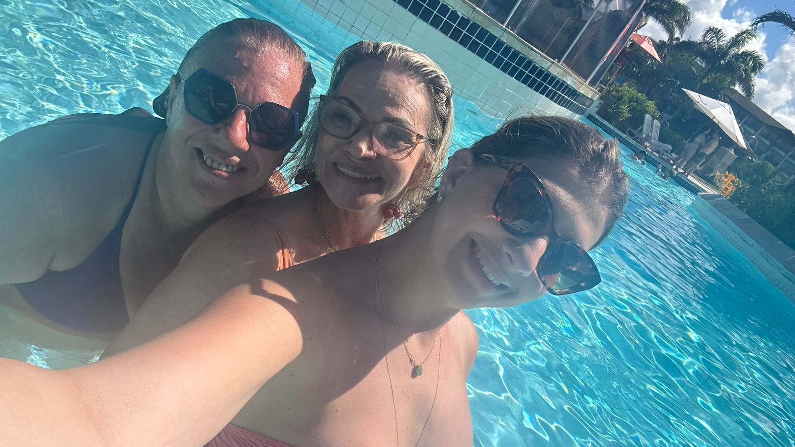 Selfie próxima de três amigas sorrindo na piscina.