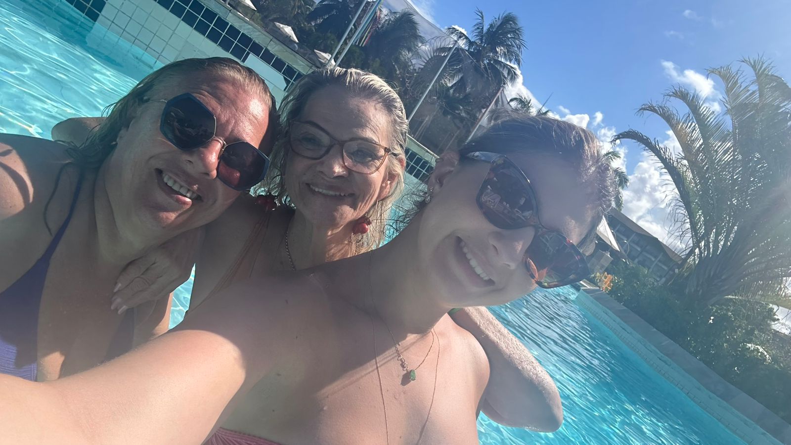 Trio junto na borda da piscina, céu azul ao fundo.