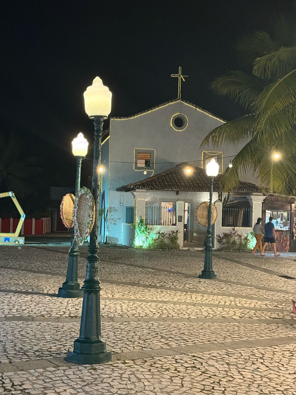 Praça com postes antigos e igrejinha iluminada à noite.
