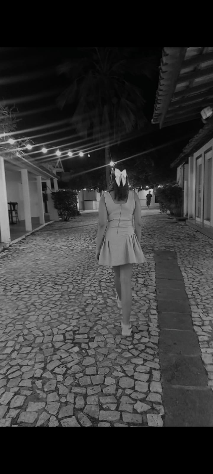 Silhueta caminhando por corredor noturno com laço no cabelo, luzes ao fundo.